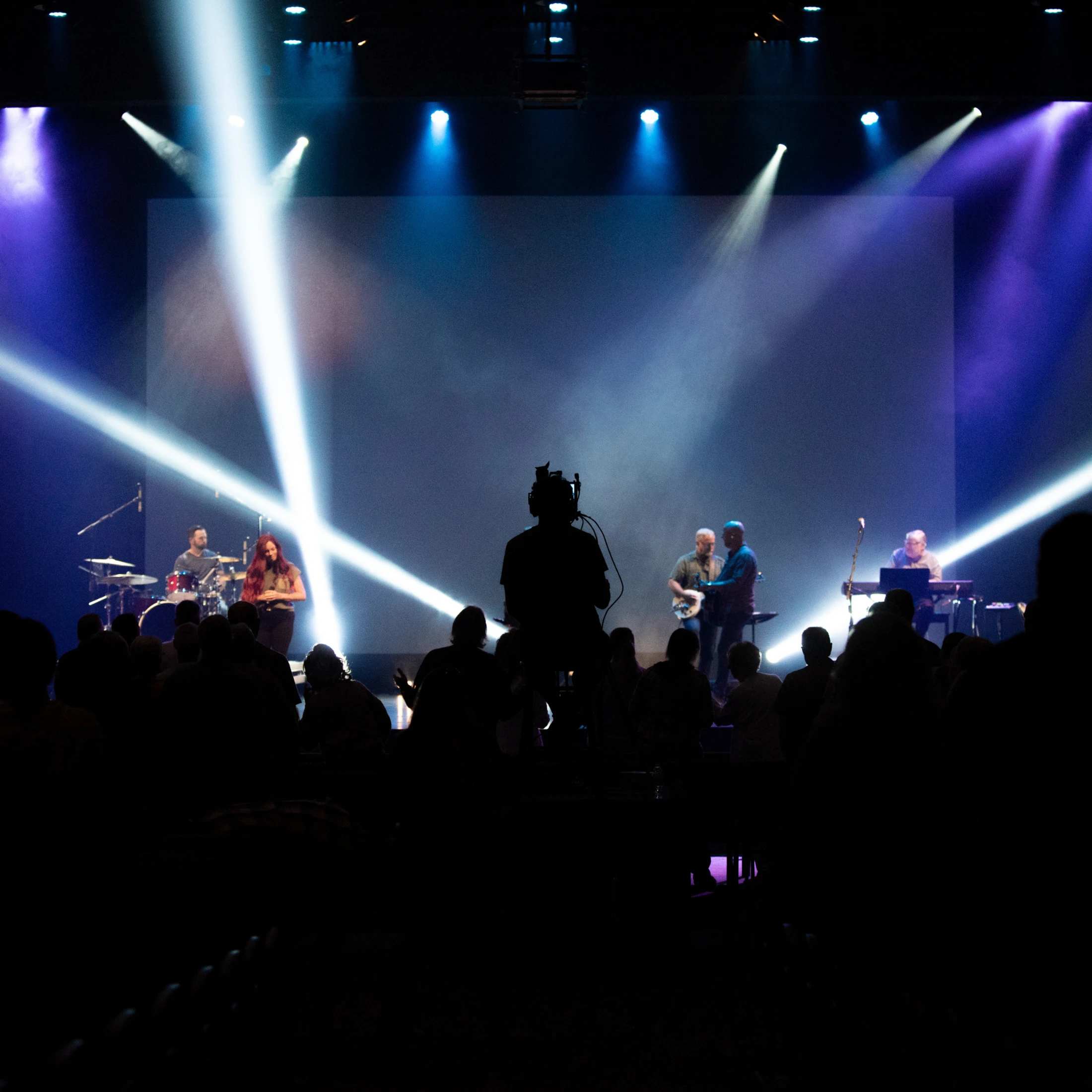 View from behind a silhouetted audience and camera operator as a band performs on a stage lit by dramatic white and blue spotlights.